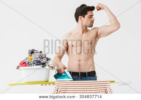 Handsome brunette shirtless houseman wearing apron standing isolated over white background, ironing clothes on a board, flexing muscles