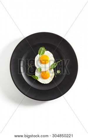 Elegant Restaurant Plate of Fried Eggs Decorated with Fresh Spinach Leaves and Young Pea Shoots on Flat Black Plate. Macro Photo of Healthy Nutritious Breakfast Isolated on White Background
