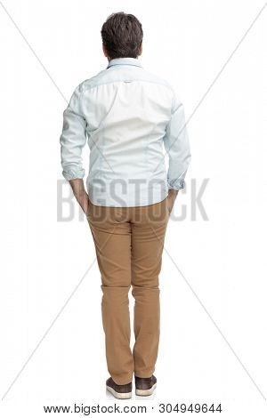 Rear view of casual old man standing with his hand in his pockets while wearing brown pants and a white shirt on white studio background