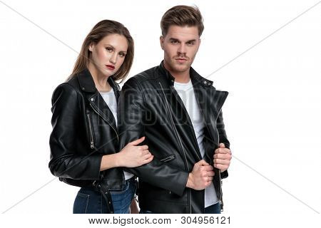 Motivated young man pulling his jacket and being embraced by his girlfriend, both wearing black leather jackets and white t-shirts, standing on white studio background