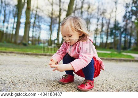Adorable Baby Girl In Park, Sitting On Hunkers And Playing With Little Stones. Little Child Having F