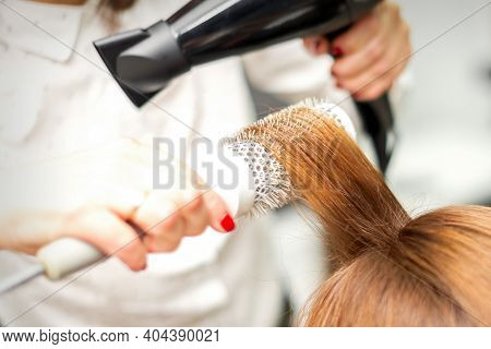 Close Up Of Hairdresser Drying Long Red Hair With A Hairdryer And Round Brush In A Beauty Salon