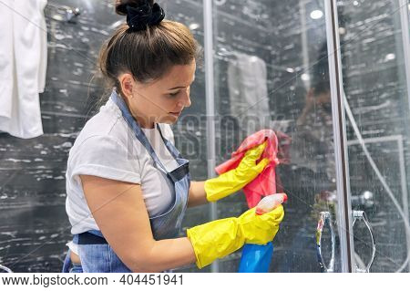 Cleaning Lady In Apron Gloves With Detergent Washing Glass Of Shower Cabin In Bathroom, House Cleani