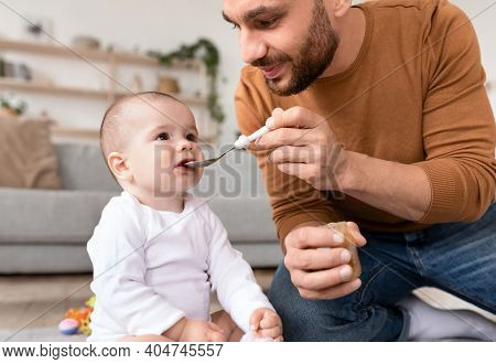 Loving Young Father Spoon Feeding His Little Baby Daughter Caring For Toddler During Paternity Leave