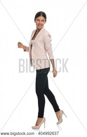 side view of beautiful young woman in pink jacket smiling and looking over shoulder, walking isolated on white background in studio, full body