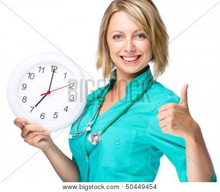 Young happy lady doctor is holding clock showing seven, isolated over white