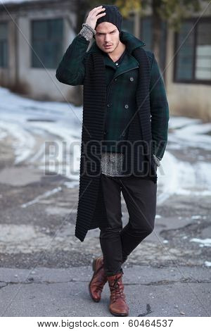 full length photo of a young fashion man posing outdoor with a hand on his cap and one in his pocket while looking into the camera
