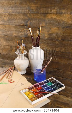 Composition with colorful watercolors, brushes in vase and sketcher on wooden background 