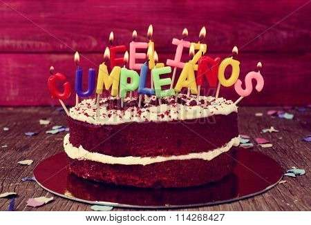 a cake topped with some lit letter-shaped candles forming the text feliz cumpleanos, happy birthday in Spanish, before blowing out the cake, on a rustic wooden table