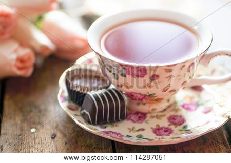 Close Up Of A Cup Of Tea With Roses And Chocolate Candies On Wooden Table
