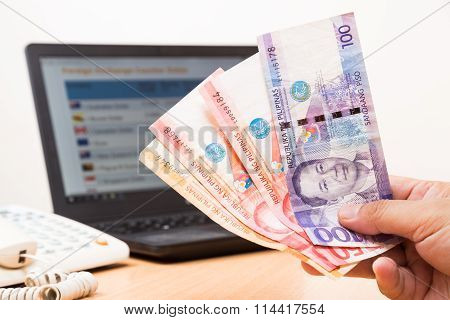 Hand Holding Philippines Piso In Office With Computer In Background