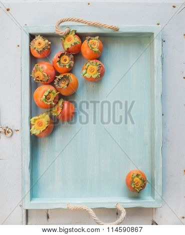 Ripe fresh persimmons on blue wooden tray over light backdrop, top view