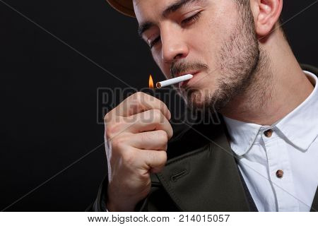 A man on a black background, retro style, in a cap lights a cigarette, screwing up his eyes.