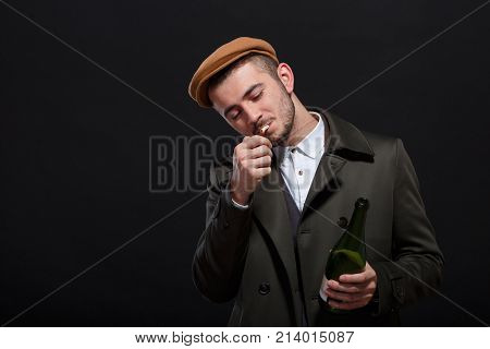 A man on a black background, retro style, in a cap lights a cigarette, screwing up his eyes, holding a bottle of alcohol in his hand.