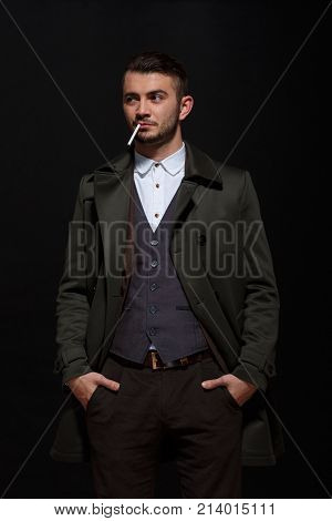 A man on a black background, retro style, stands with a cigarette in his mouth, putting his hands in his pockets, looking away.
