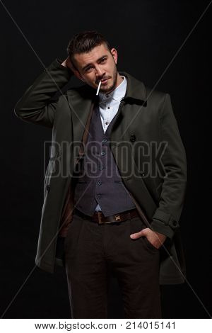 A man on a black background standing with a cigarette in his mouth, scratches his head with his hand, putting hand in pocket, looking at the camera.