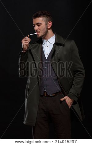 A man on a black background, standing with a cigarette in his mouth, lights a cigarette, putting one hand in his pocket