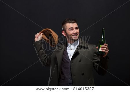 A man on a black background, retro style, holds in his hand a bottle of alcohol, in the second hand a brown cap, which he tries to throw away.