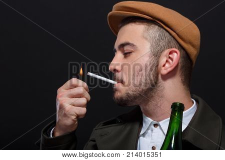 A man on a black background, retro style, in a cap lights a cigarette, screwing up his eyes, holding a bottle of alcohol in his hand.