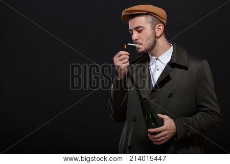 A man on a black background, retro style, in a cap lights a cigarette, screwing up his eyes, holding a bottle of alcohol in his hand.