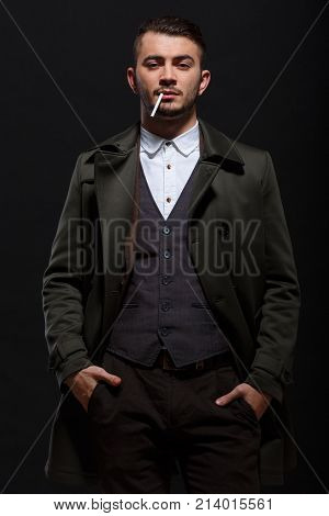 A man on a black background, retro style, stands with a cigarette in his mouth, putting his hands in his pockets, looking away.