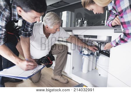 A woman shows plumbers a broken kitchen sink. One of the men inspects the pipes, the second writes information to the form. A woman is watching the work.
