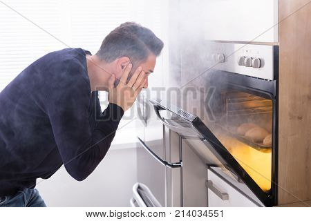 Shocked Man Looking At Burnt Cookies With Smoke Coming From Oven