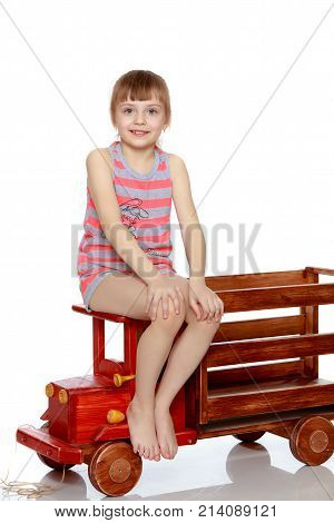 Beautiful little caucasian girl with a short bangs and hair tucked into a tail. In striped in a T-shirt and shorts.The girl is sitting on a large toy wooden car.
