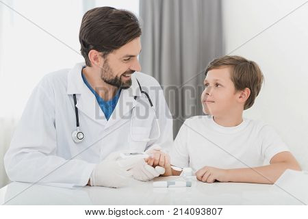 The doctor takes the blood from the boy with a finger using a scarifier. The boy staunchly endures the procedure. The doctor will check the boy's blood for sugar level.