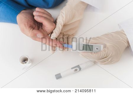 A nurse takes a blood sample with an old man's scarifier. The nurse takes the blood to the glycemeter. They are sitting at a white table.