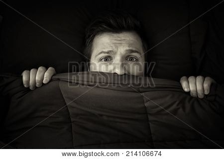 Caucasian young man hiding in bed under the blanket at home. He is stressed and tries run away from problems. Black and white photo