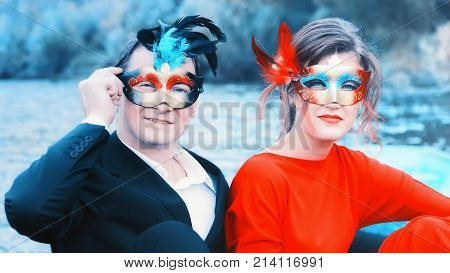 Colorful portrait of a young enamored couple in masquerade masks - man in a suit and woman in red against a blue lake. Focus on foreground.