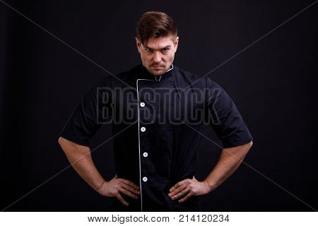 A man pumped up in a black tunic with a serious and confident gaze holds his hands in the sides on a black background