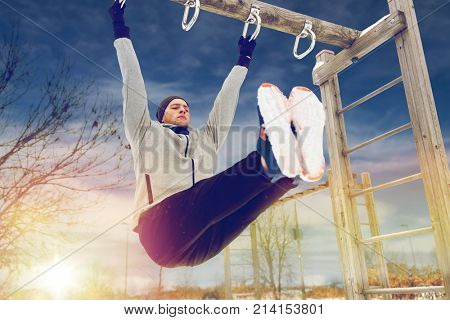 fitness, sport, exercising, training and people concept - young man doing leg pull ups on horizontal bar and flexing abdominal muscles outdoors in winter