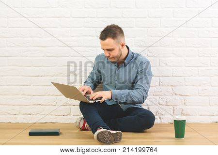 Attractive guy sitting in studio with laptop book and coffee cup on wooden floor. White brick wall background. Education youth hipster concet