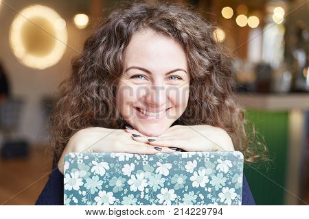 Youth and happiness. Portrait of young Caucasian female wearing elegant blue sweater using free wireless internet connection on laptop in original cover waiting for order at cafe smiling at camera.