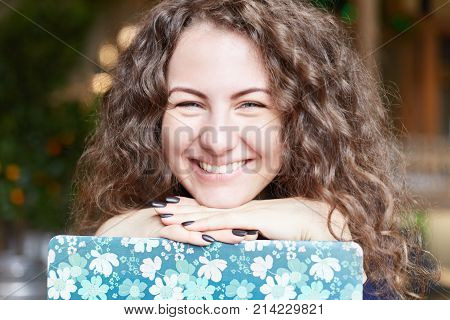 Horizontal portrait of adorable lady dressed casually has happy expression demonstrates white teeth as smiles sits over cafe interior with opened laptop in colorful cover waits for companion.