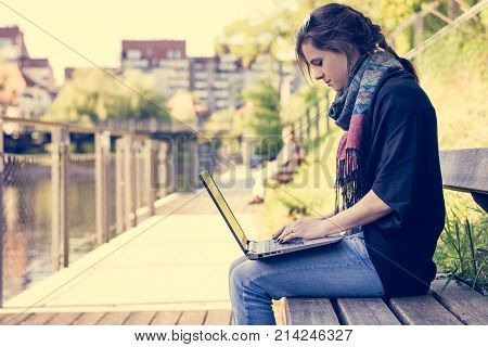 Young woman using laptop at a riverbank. Milleanial working in a park.