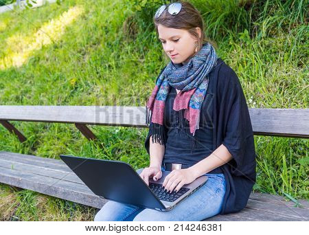 Young woman using laptop at a riverbank. Milleanial working in a park.