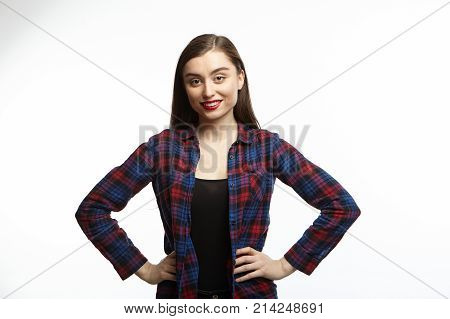 Studio shot of attractive self confident young female in great mood feeling happy holding hands on her slender waist and looking at camera with radiant smile rejoicing at good day or news