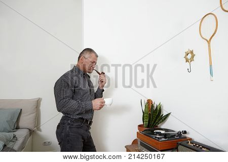 Stylish senior man wearing shirt and jeans standing in modern apratment interior hoding cup of coffee smoking tobacco pipe and listening to music on retro vinyl record player deep in thoughts