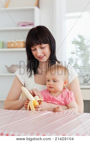 Krásná bruneta žena žáčkům banán současně držet své dítě na kolenou v kuchyni