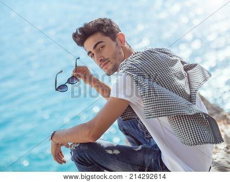 Traveler on the rocks near the sea looking at camera. Summer Travel Vacation. Smiling handsome young caucasian tourist man in casual clothes outdoors on the nature.