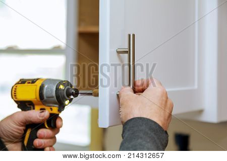 The worker sets a new handle on the white cabinet with a screwdriver installing kitchen cabinets