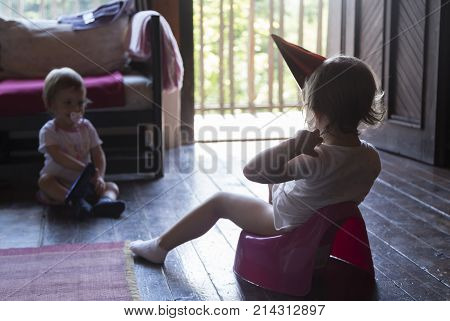 little baby sitting on a potty with party hat on the cottage with his younger sister