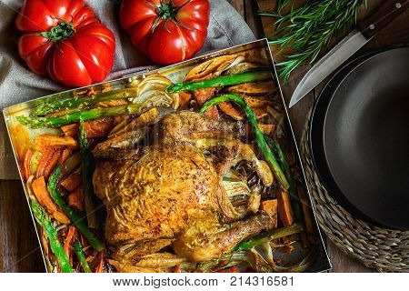 Homemade Roasted Stuffed Chicken Vegetables in Baking Form Yams Carrots Asparagus Onions Herbs Rosemary Thyme. Festive Dinner Dish Christmas Thanksgiving Table Setting Dark Plates Knife Top View