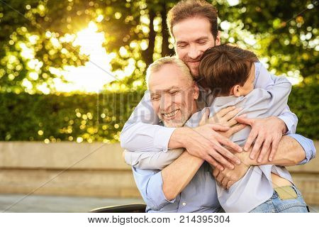 Family meeting. A man and a boy came to see his grandfather, who is sitting in a park on a wheelchair. The old man, his son and grandson embrace, they are happy