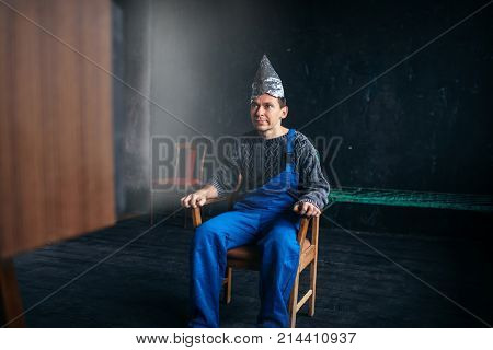 Man in tinfoil hat sits in chair, paranoia concept