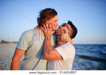 Two men embrace against the background of the sea. They represent a playful kiss.