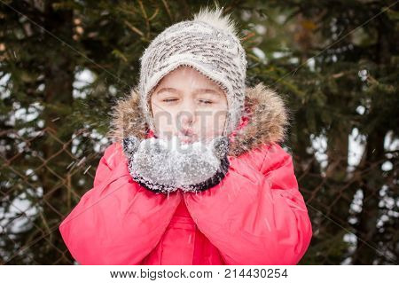 Happy Child Portrait Of Little Girl With Eyes Closed In Red Jacket Blowing Fluffy Snow Out Her Hands In Winter Park Outdoor Close Up. Playing With Snow.